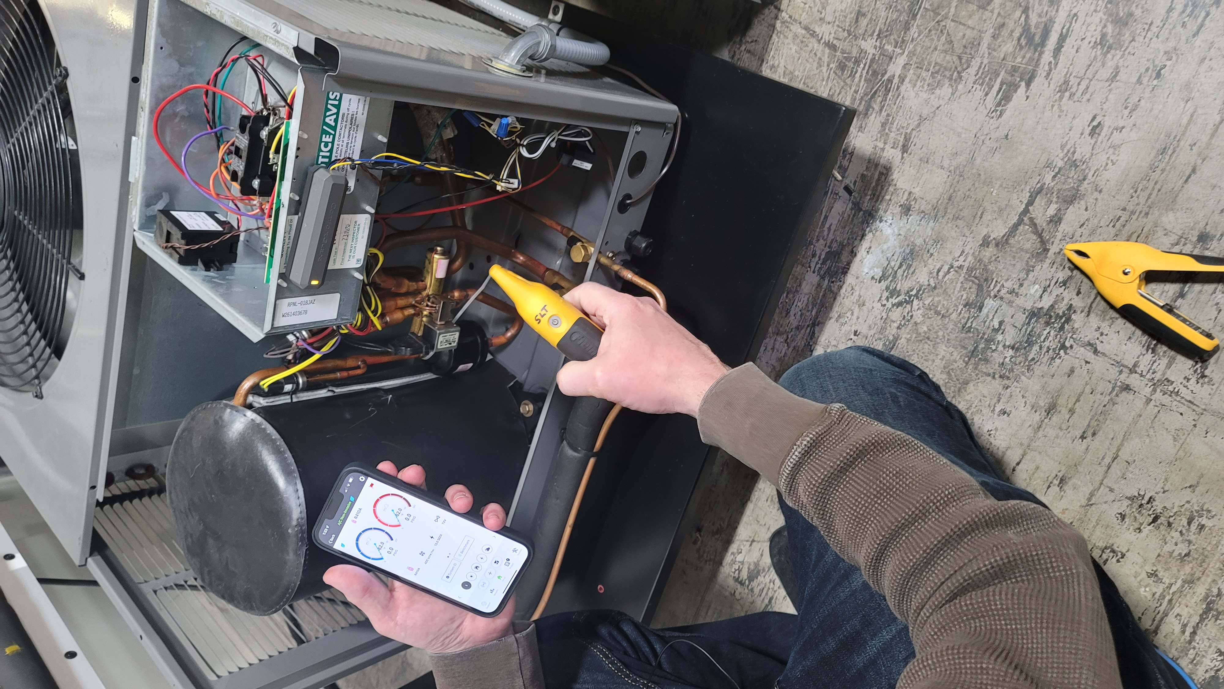 Technician at outdoor unit with panel removed, placing pipe clamp probes on the refrigerant lines while viewing measureQuick on a phone